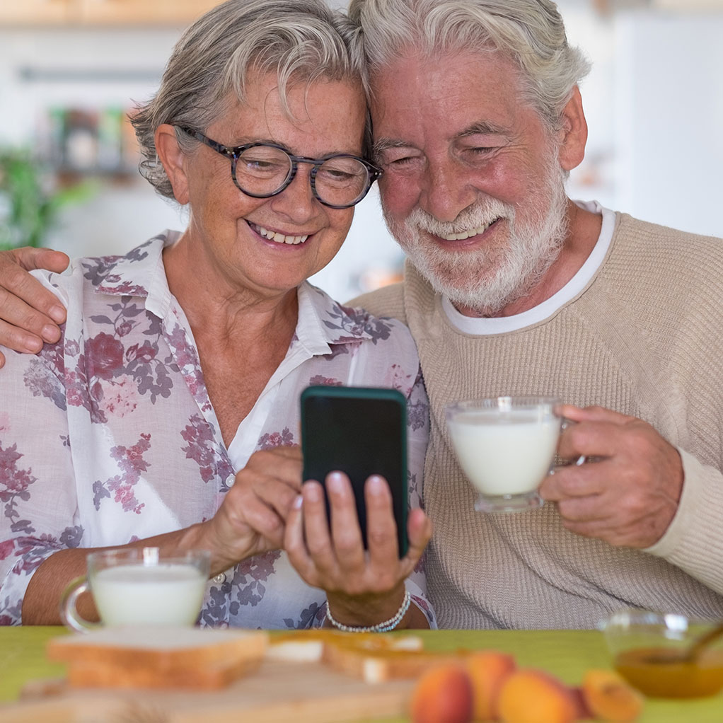 elderly couple smiling happily looking at photos on a mobile phone drinking a glass of milk each